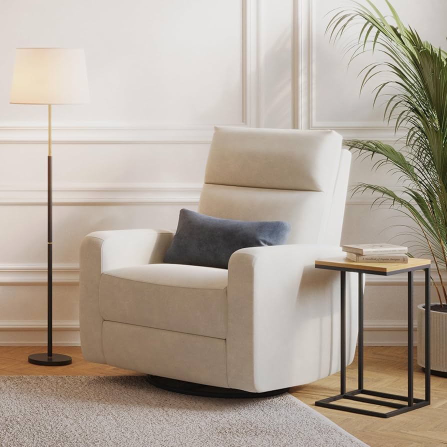 A modern cream-colored nursery glider with a blue lumbar pillow, shown in a bright, cozy room beside a small side table and floor lamp.