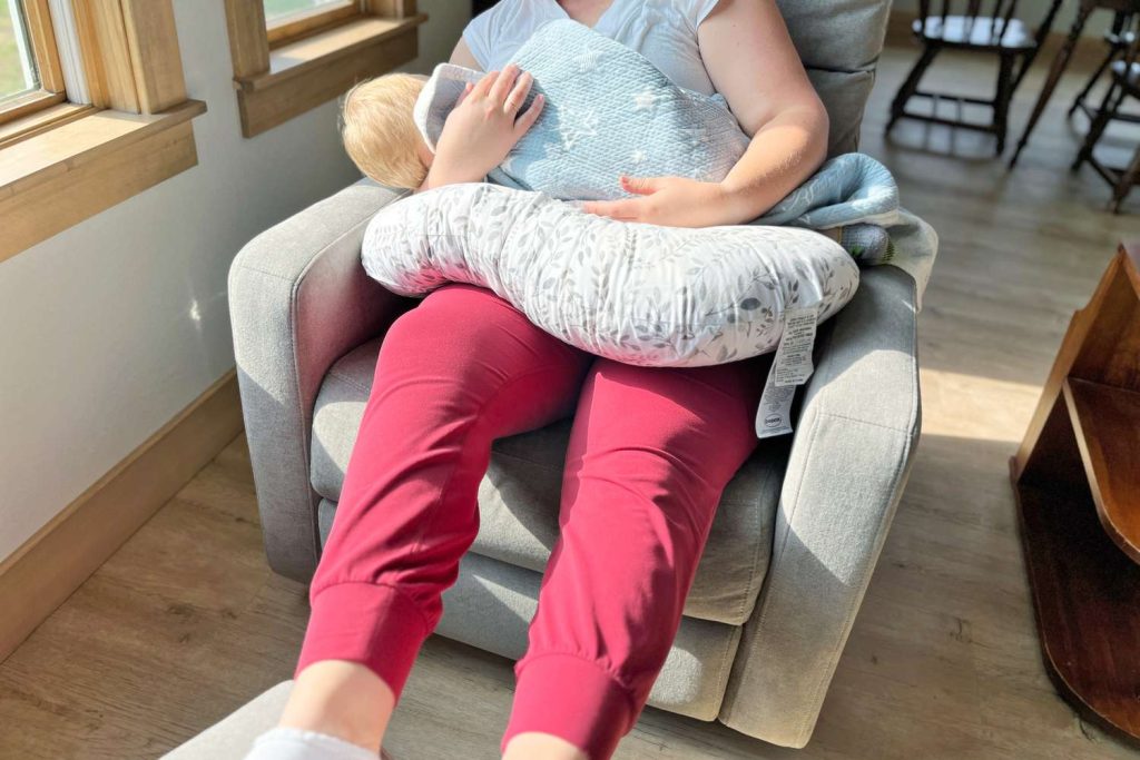 A mother breastfeeding her baby while sitting in a gray nursery glider, showing extended leg space suitable for tall parents.